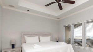 Quarto com cama, mesas de cabeceira, ventilador de teto e janelas com persianas.