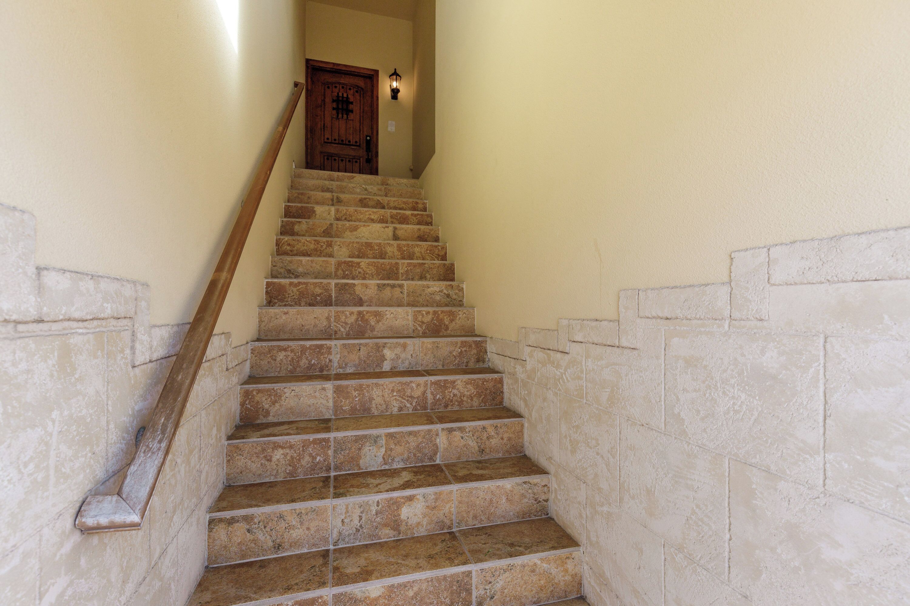 Un escalier avec des marches en pierre et une rampe en bois, menant à une porte sur laquelle est fixée une applique lumineuse.