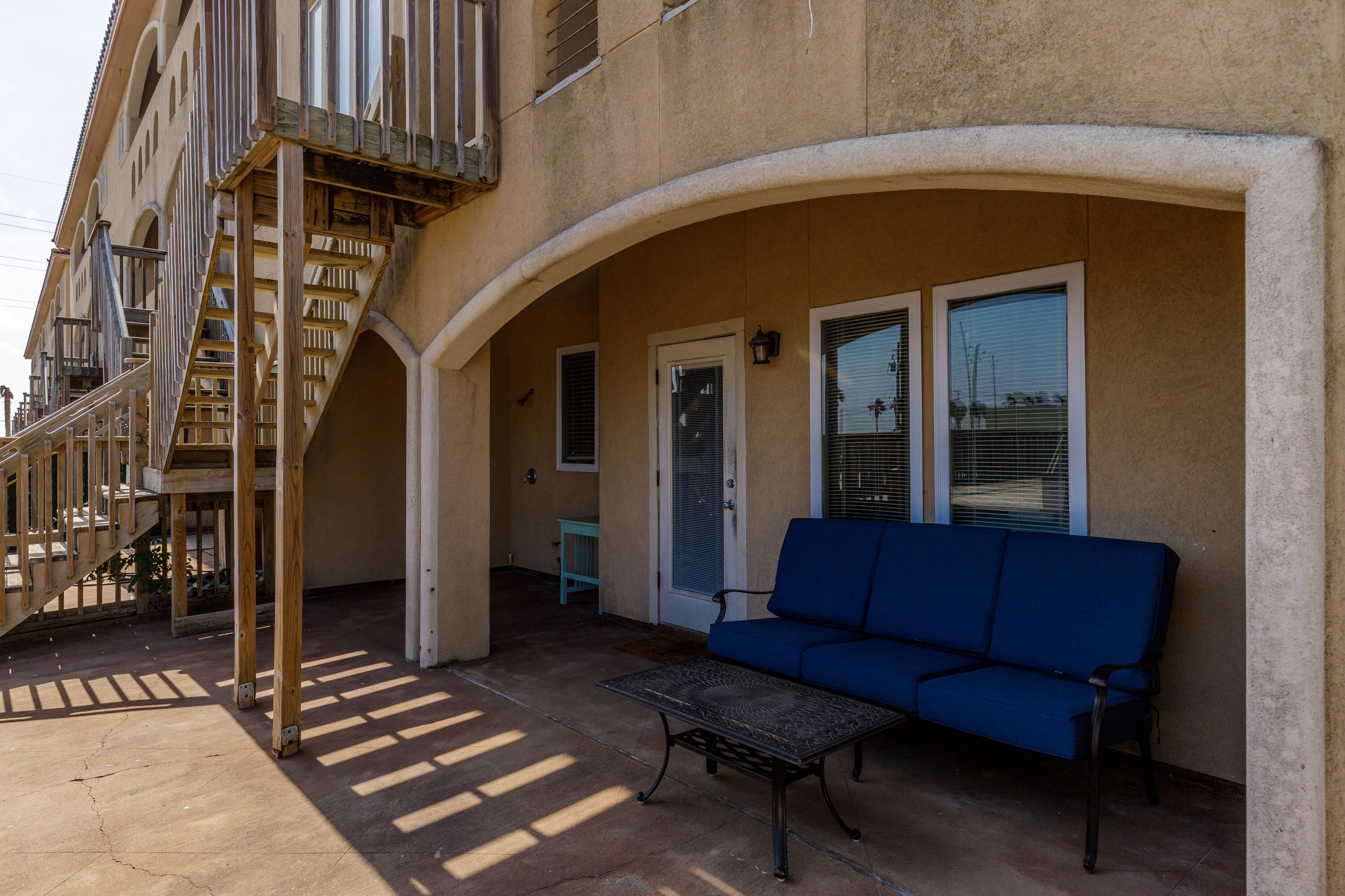 A covered outdoor area with a blue sofa, a metal table, and a staircase leading to an upper level.