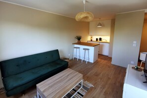 A modern living room with a green sofa, wooden coffee table, and a kitchen area with stools and a hanging light fixture.
