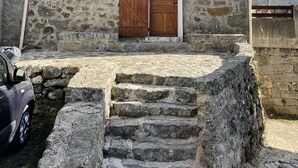 Ein Steinhaus mit einer Holztür und -läden, einem kleinen Fenster und einer Treppe aus Stein, die zum Eingang führt.