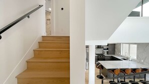 A staircase with wooden steps and a metal handrail, leading to a modern kitchen with a marble countertop and a dining area with a table and chairs.