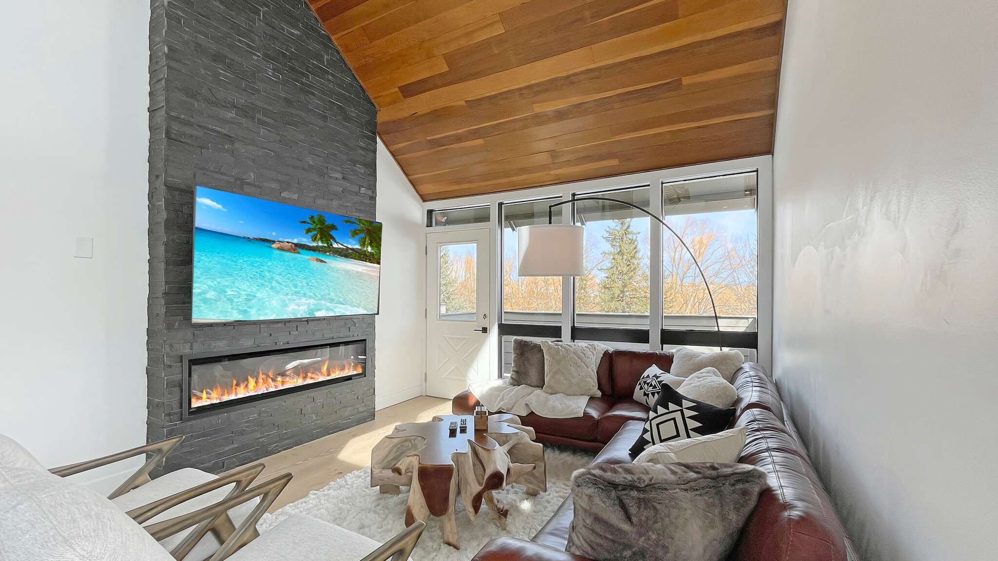 Ein Wohnzimmer mit Kamin, einem Fernseher der eine Strandansicht zeigt, und einem großen Fenster mit Blick auf Bäume.