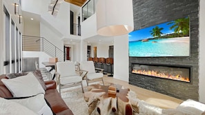 A modern living room with a fireplace, a large TV screen, and a staircase leading to an upper level.