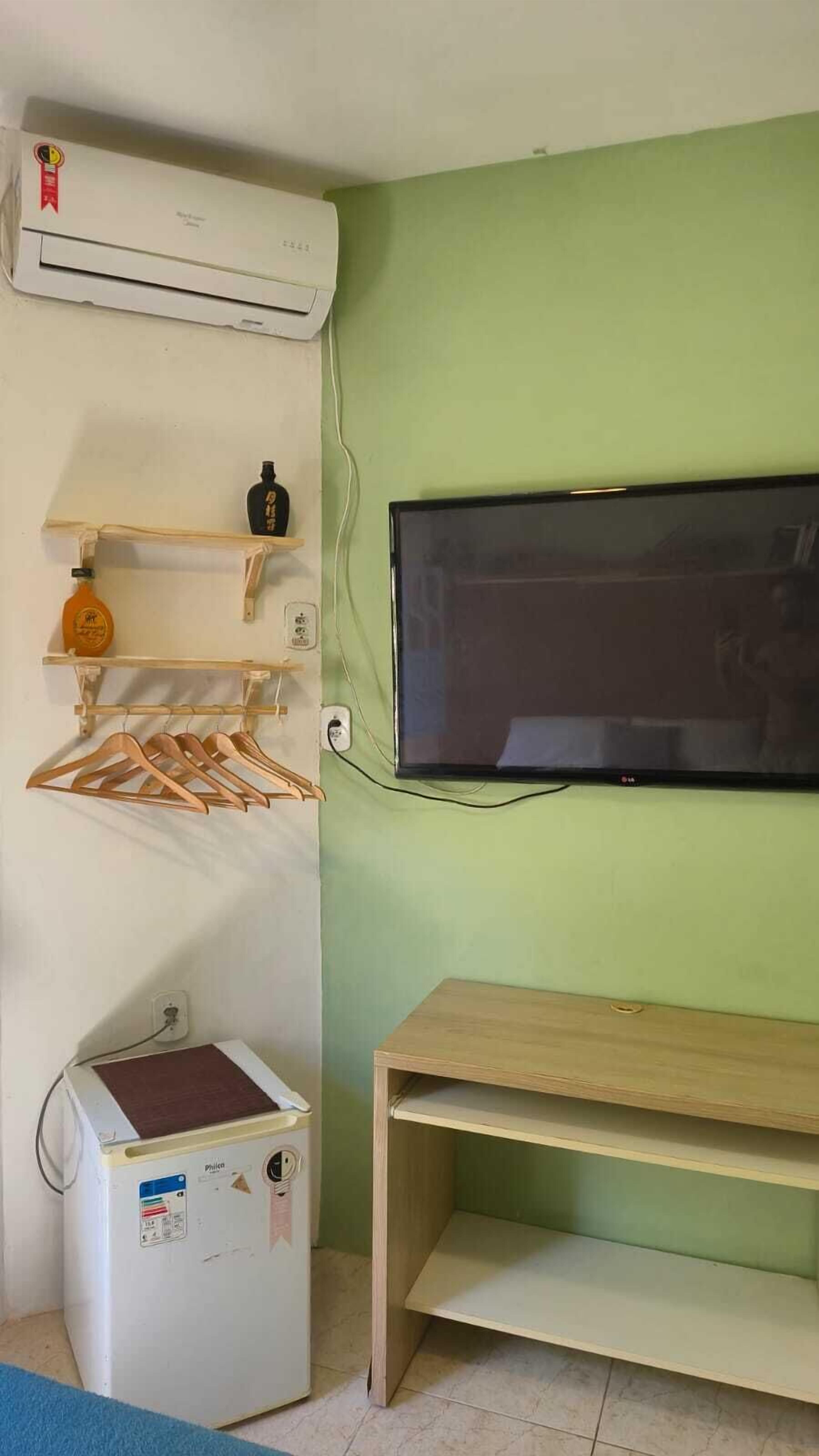 Une pièce avec un téléviseur fixé au mur, une petite étagère en bois sur laquelle se trouve une bouteille, un porte-manteau en bois, un appareil électroménager blanc doté d’un panneau de commande et un banc en bois.