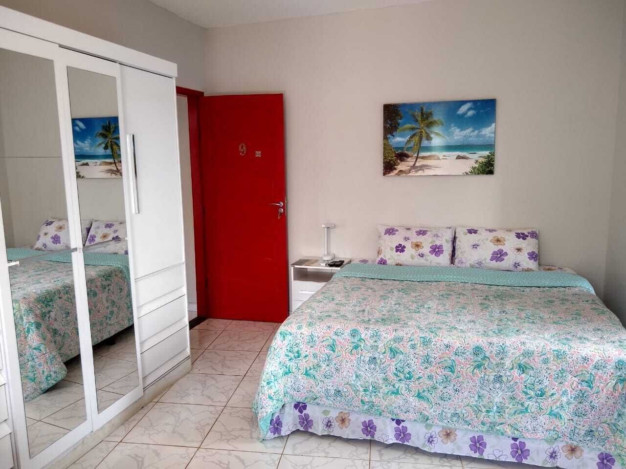 A bedroom with a bed, a red door, a nightstand, a painting of a beach, and a wardrobe.