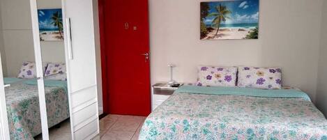 A bedroom with a bed, a red door, a nightstand, a painting of a beach, and a wardrobe.