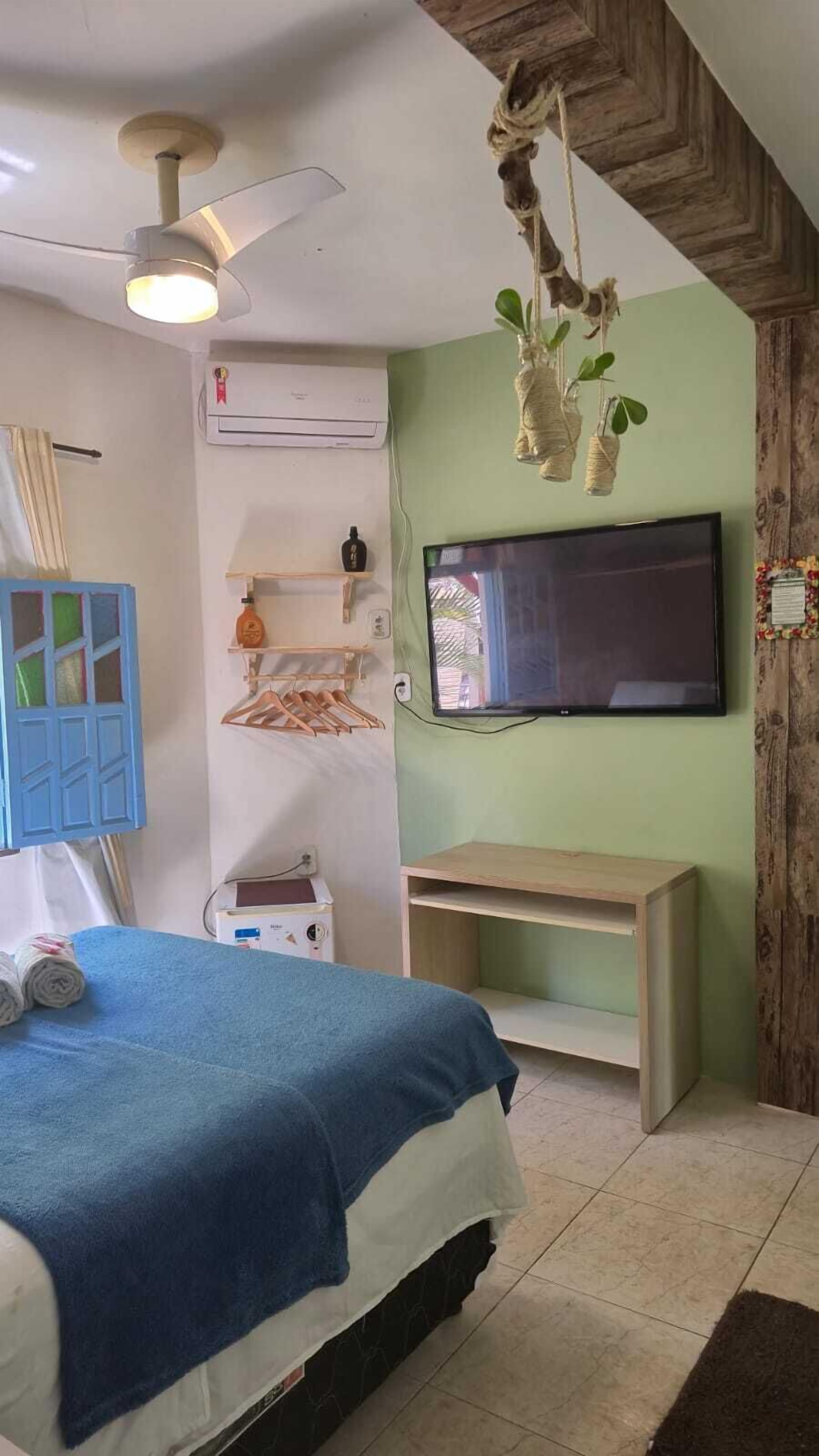 Quarto com cama, ventoinha no teto, ar condicionado, televisão e prateleira de madeira.