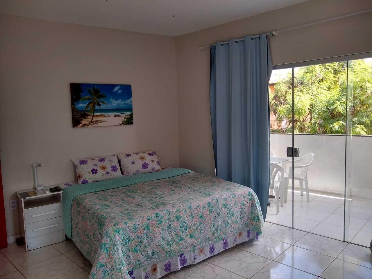 Una camera da letto con un letto, un comodino, un quadro, una finestra con tende e vista sul verde.