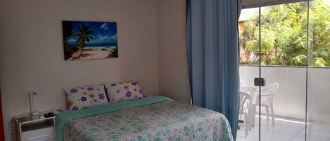 Una camera da letto con un letto, un comodino, un quadro, una finestra con tende e vista sul verde.