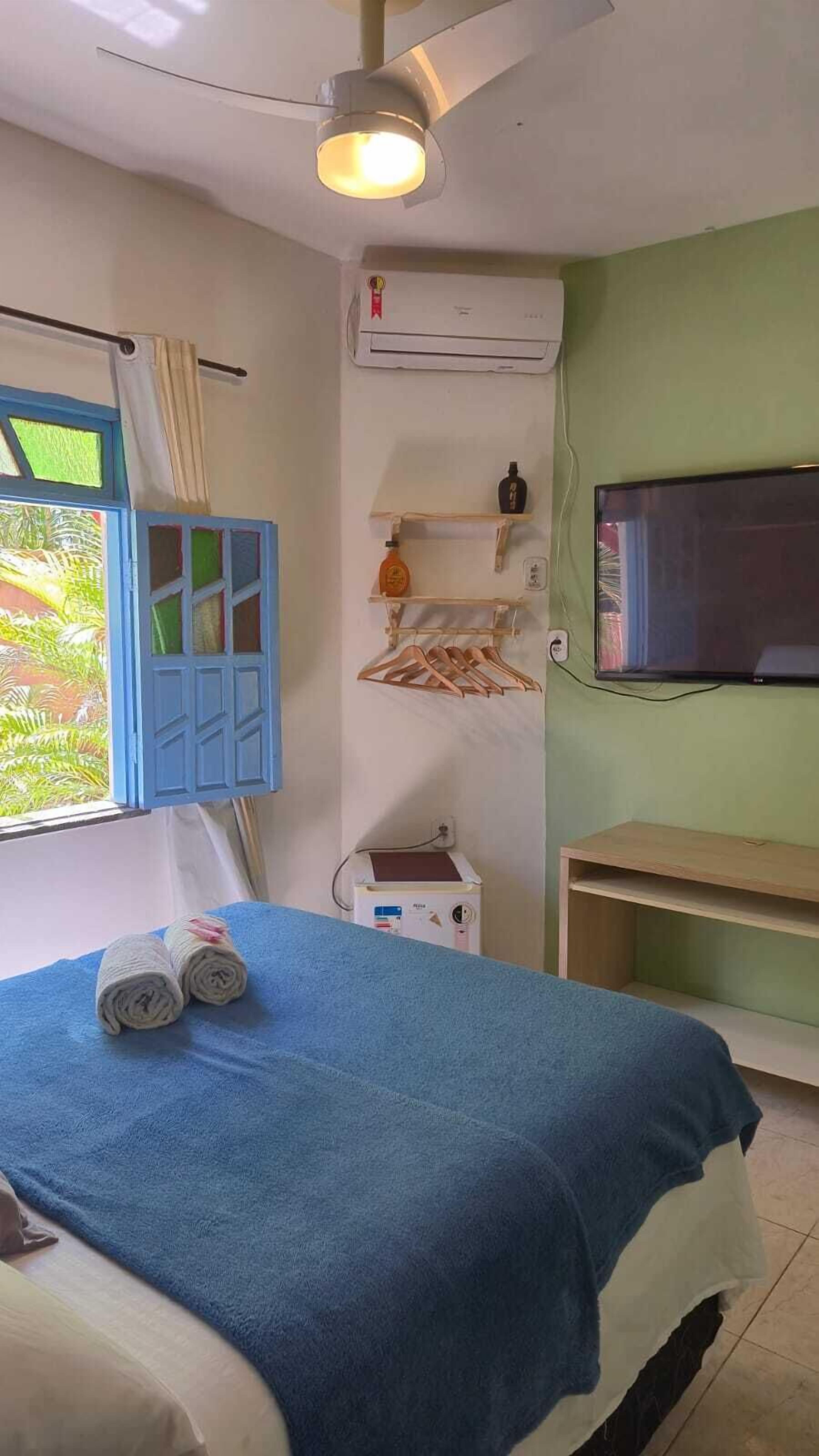 Quarto com cama, ventilador de teto, ar condicionado, televisão e janela com cortinas.