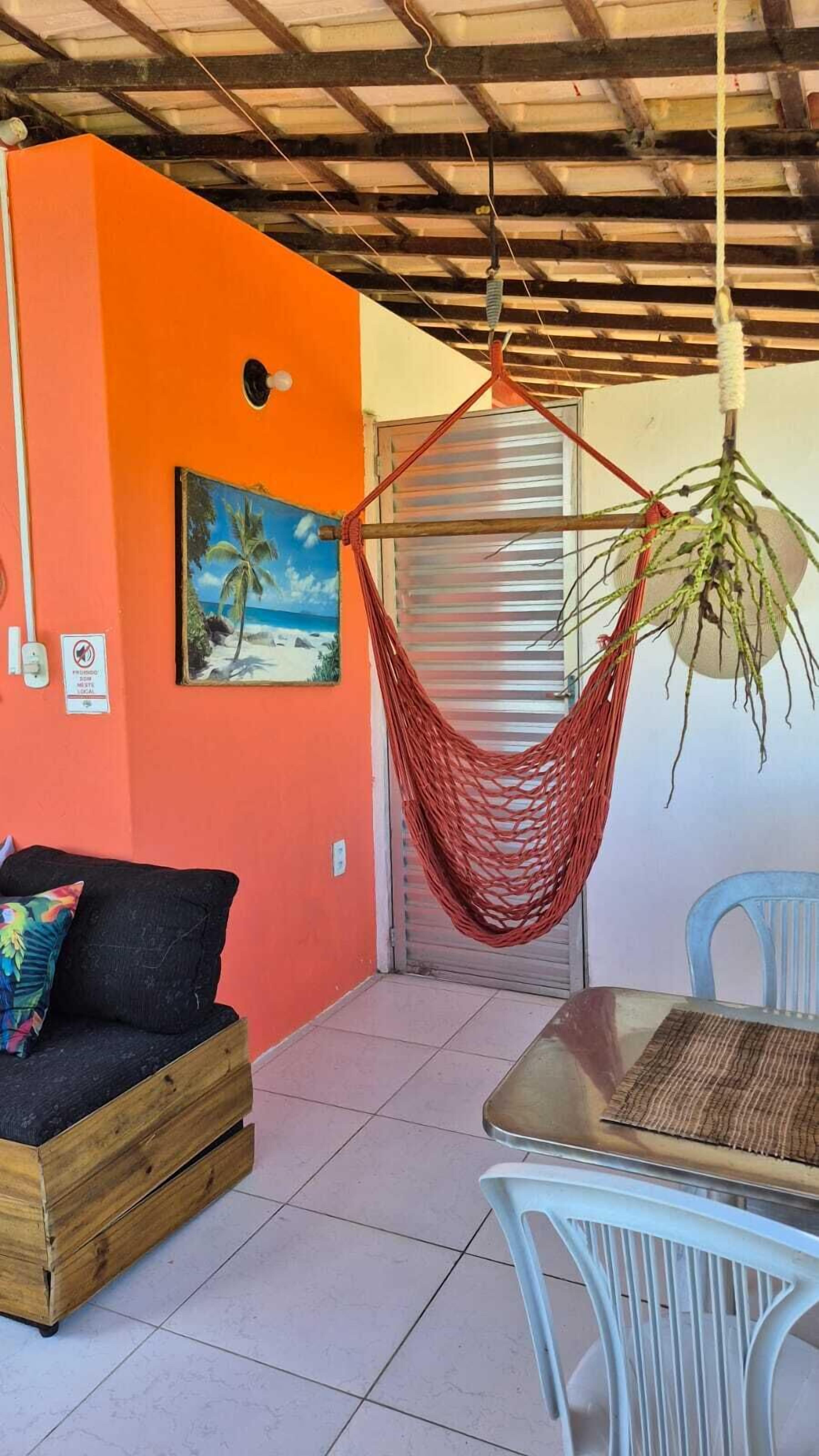 A room with a wooden ceiling, orange walls, a hammock, a beach painting, a black sofa with a colorful pillow, and a table with chairs.