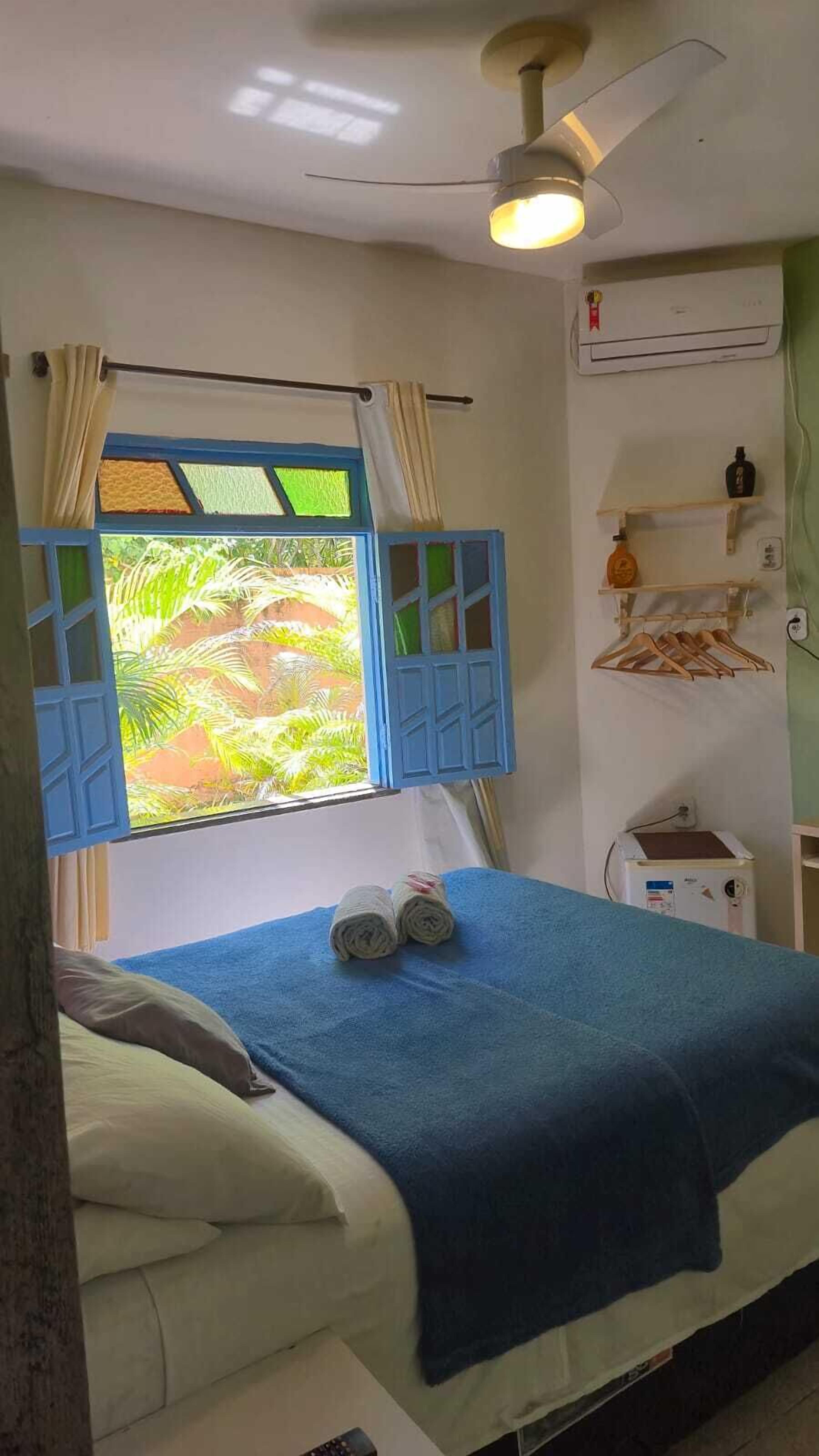 Une chambre avec un lit, une fenêtre avec des volets bleus, un ventilateur de plafond et un climatiseur fixé au mur.
