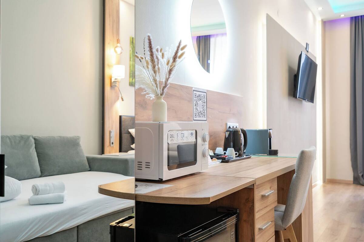Een moderne hotelkamer met een bed, een bureau met een magnetron, een stoel, een vaas met gedroogde planten, een televisie en een QR-code op de muur.