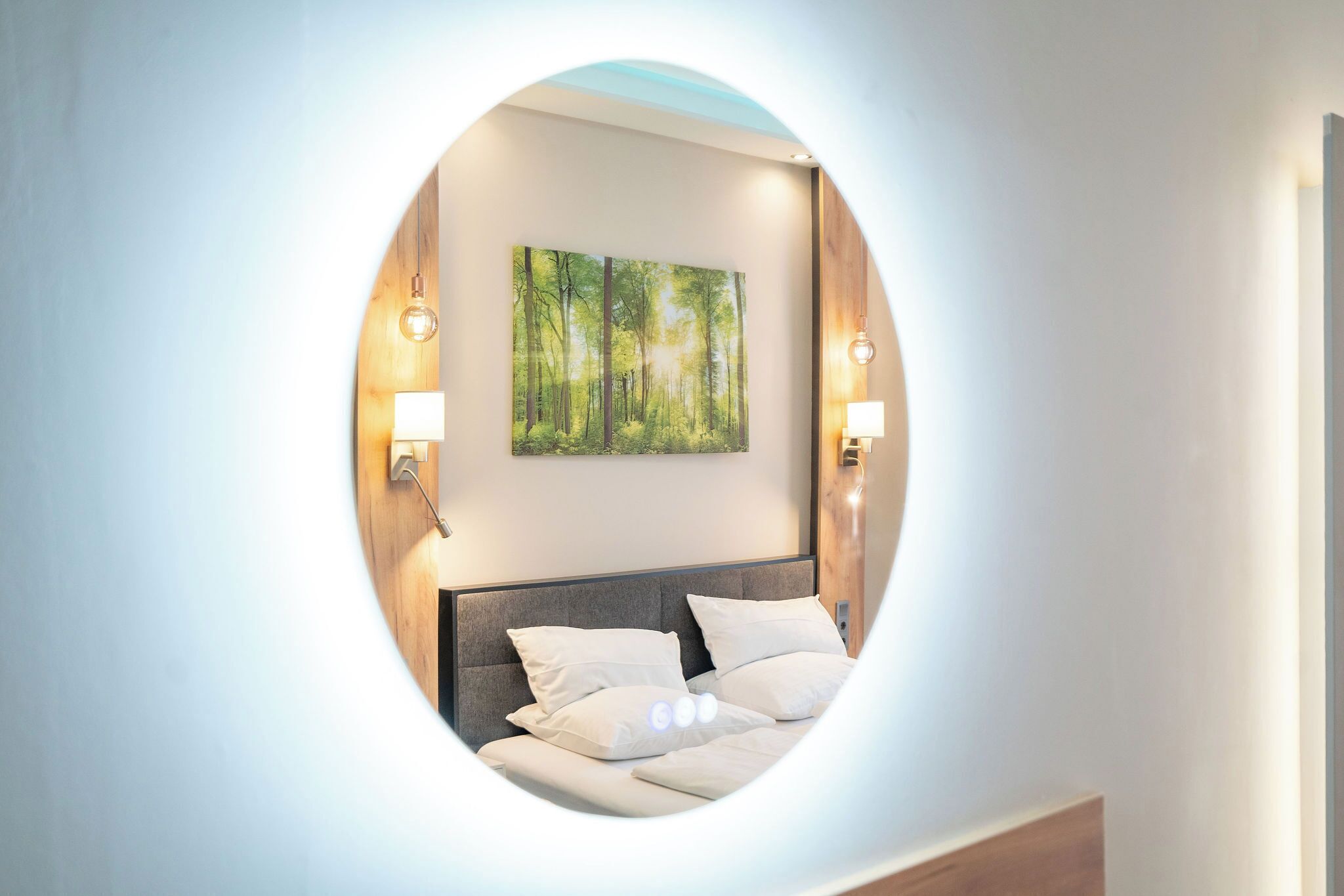 A circular mirror reflecting a bed with white bedding, a forest-themed wall art, and two wall-mounted lamps.