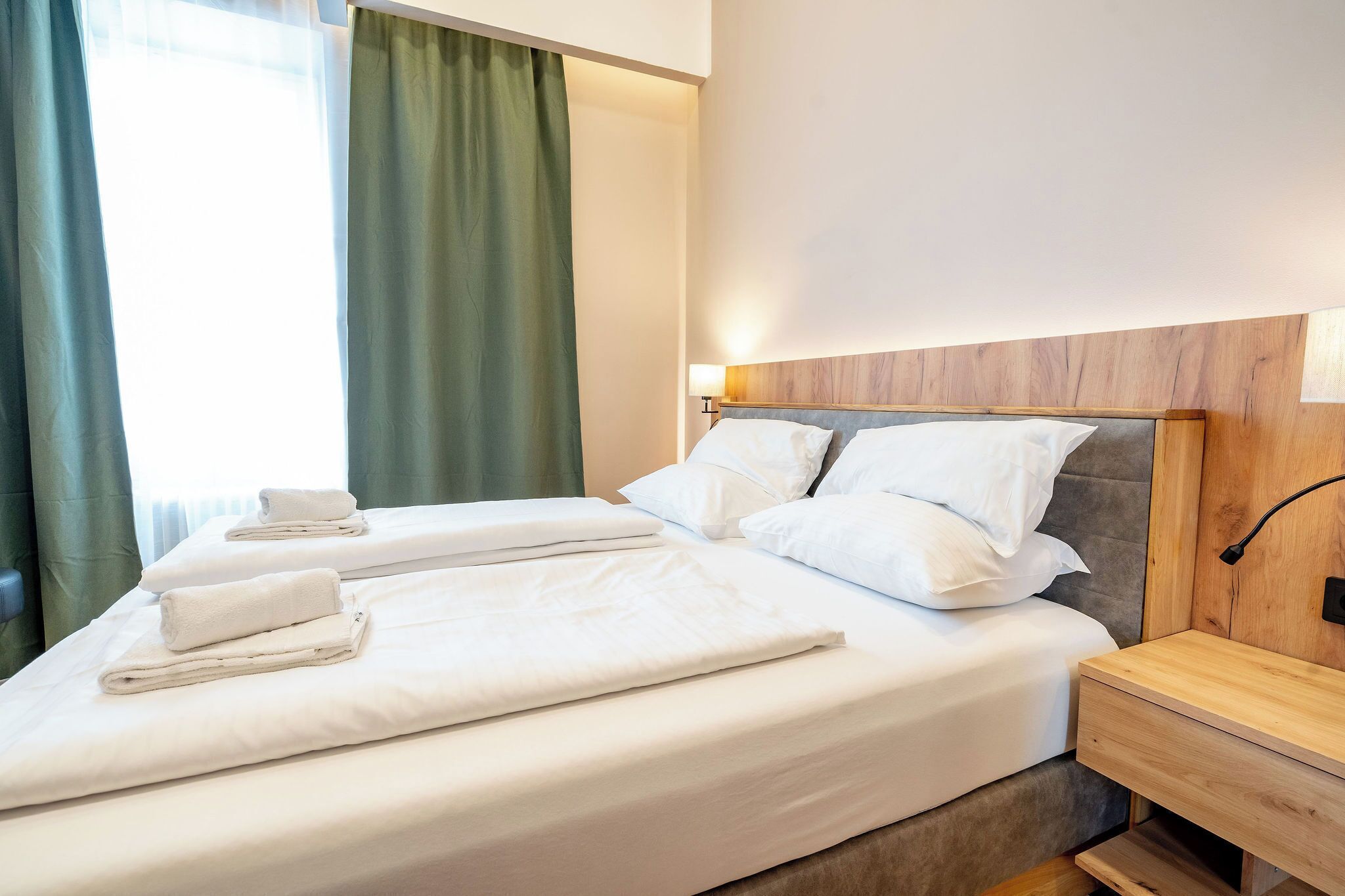 A neatly made bed with white linens and pillows, a wooden nightstand with a lamp, and a window with green curtains.