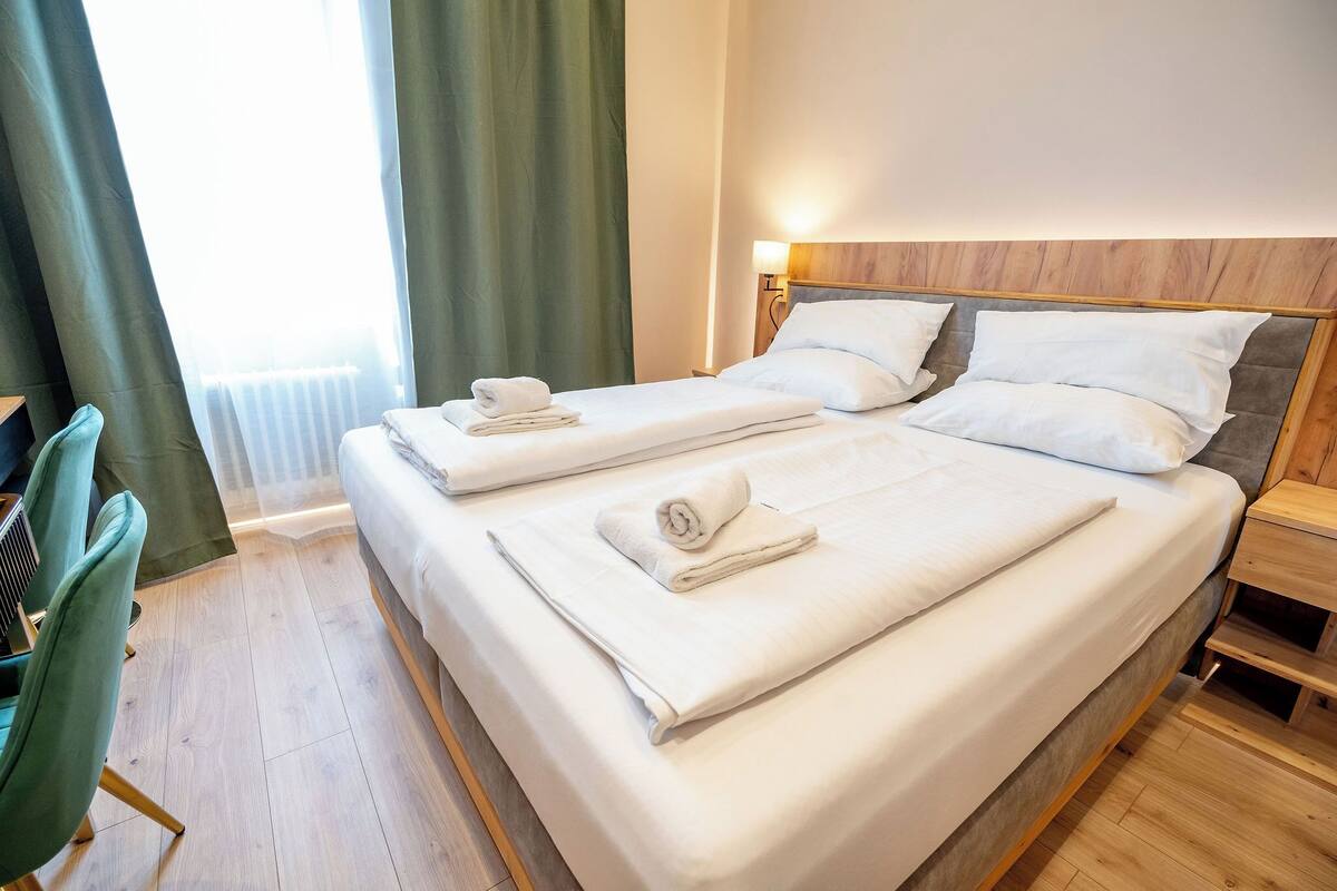 Een tweepersoonsbed met witte lakens en kussens, een houten hoofdbord, een nachtkastje en een groene stoel.