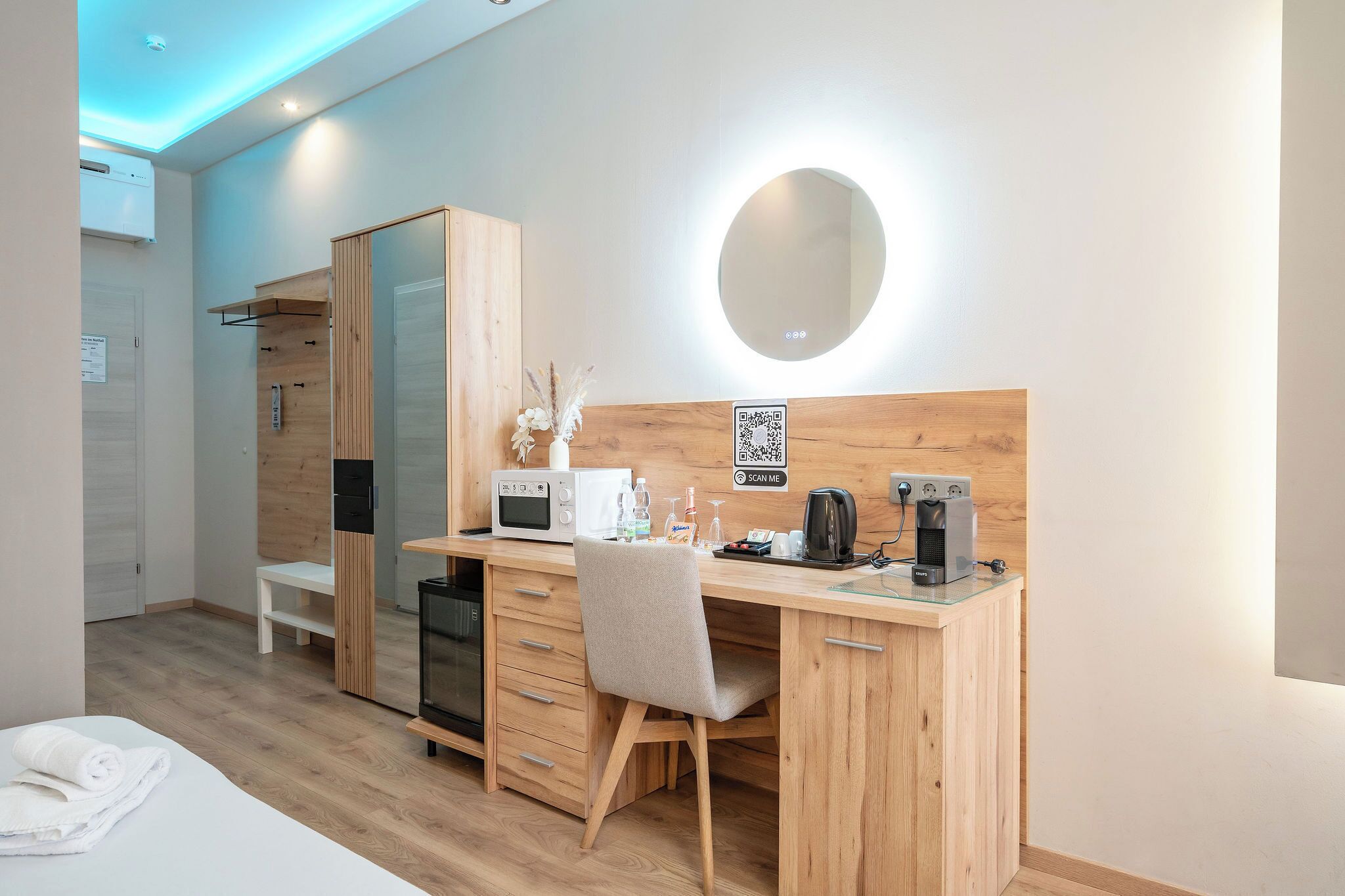 A modern hotel room with a wooden desk, a microwave, a coffee maker, a mirror, and a QR code for room service.