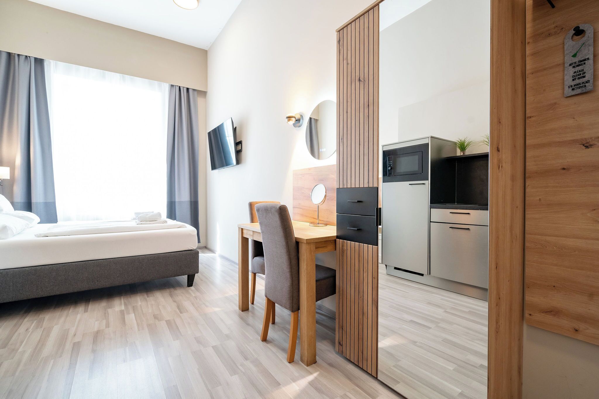 Ett modernt hotellrum med en säng, ett skrivbord med en stol, ett litet bord med en lampa, en spegel, en TV och ett litet pentry med kylskåp och mikrovågsugn.