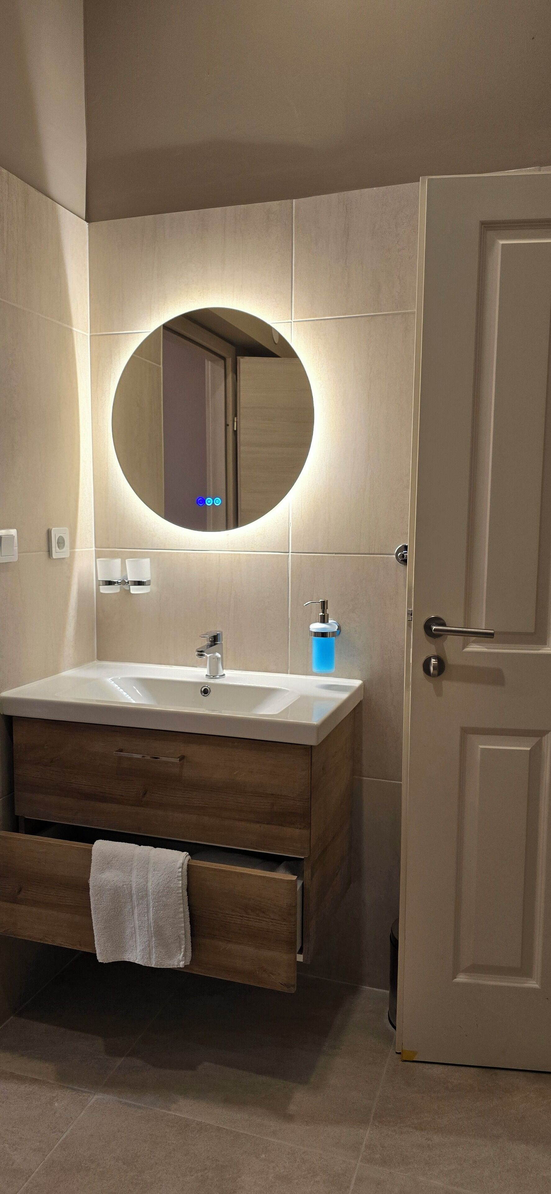 A bathroom with a wooden vanity, a round mirror, a white sink, and a door.