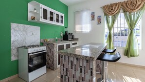 Eine Küche mit Gasherd, Mikrowelle und Backofen. Eine grüne Wand, ein Fenster mit Jalousien und eine Theke mit Hockern.