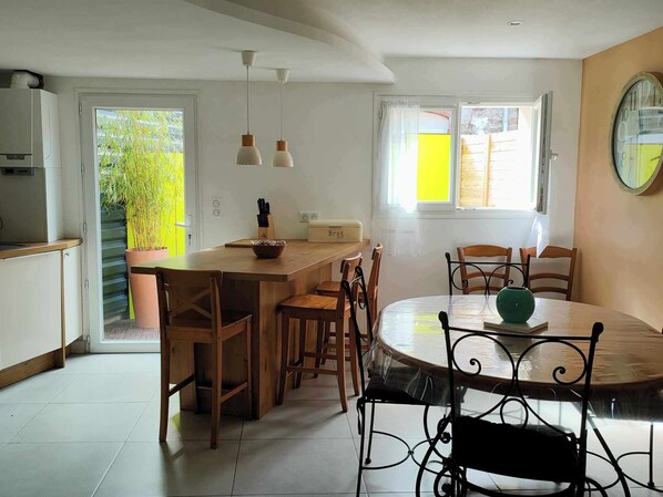 A modern kitchen with a central island, wooden chairs, a dining table with metal chairs, pendant lights, and a window with curtains.