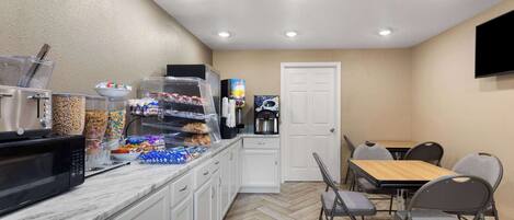 Una kitchenette con microondas, cafetera y varios recipientes de comida. Hay un comedor con mesa y sillas.