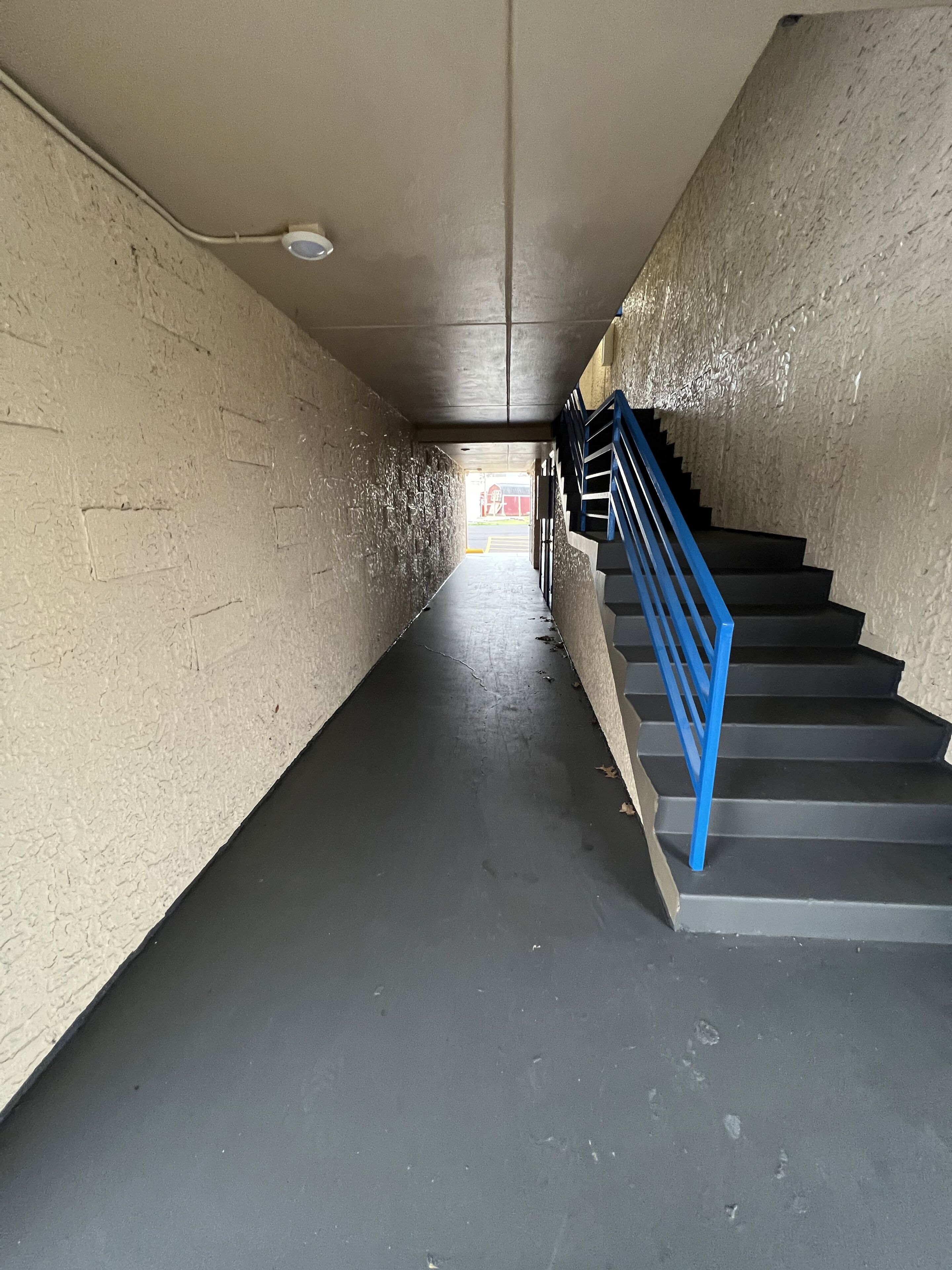 A narrow stairwell with a blue handrail, concrete walls, and a visible exit door at the end.