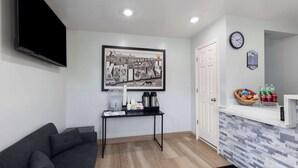 A modern living room with a sofa, a coffee table, a framed picture, a clock, and a small kitchen area with a microwave and beverages.