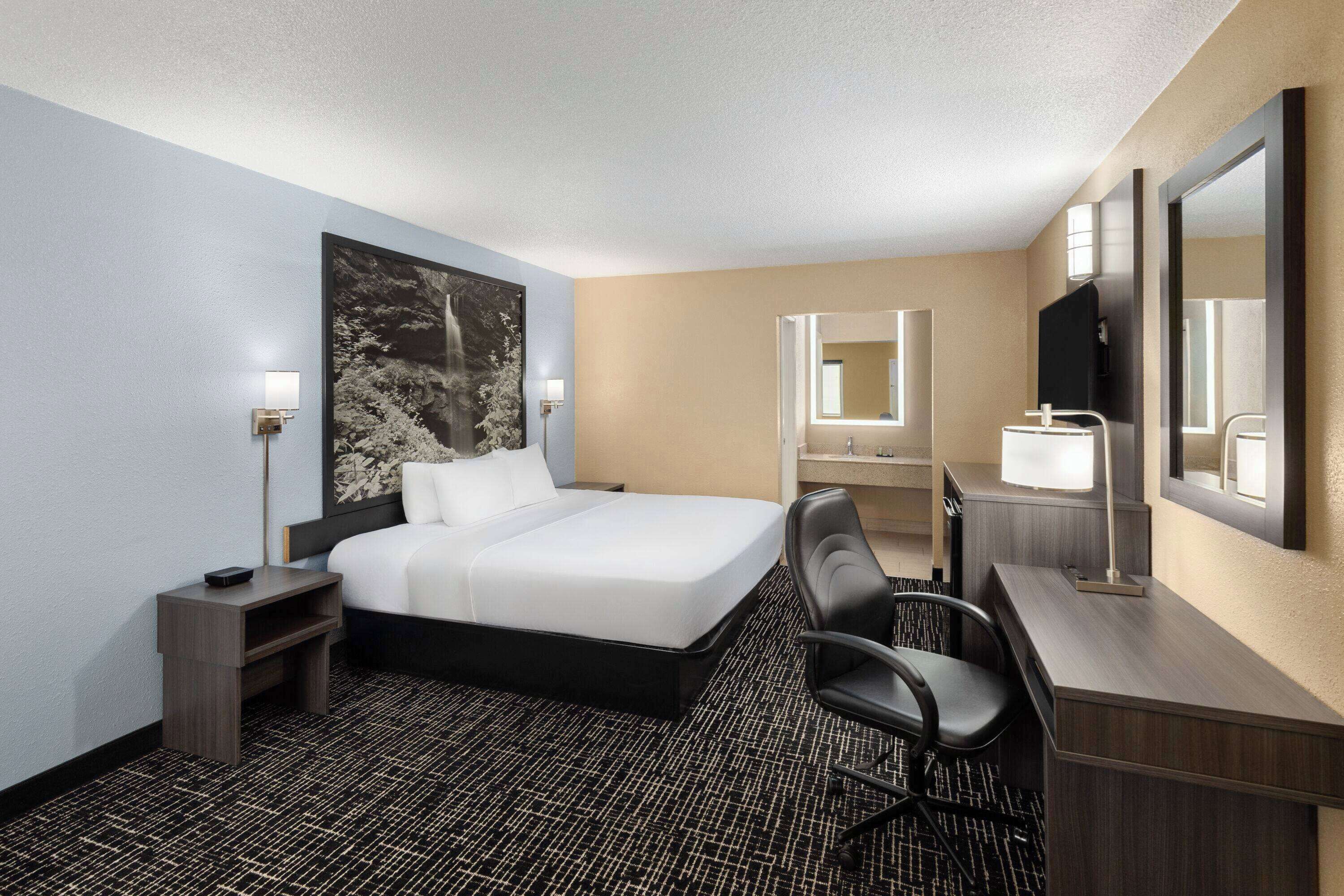 A hotel room with a large bed, a desk with a chair, a desk lamp, a flat-screen TV, a nightstand, and a wall-mounted mirror.