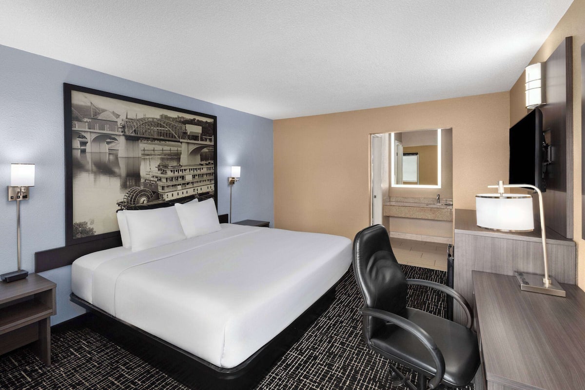 Une chambre d’hôtel avec un grand lit, un bureau, une chaise, une lampe de bureau, une table de chevet, un téléviseur fixé au mur et un tableau représentant un pont.