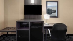 A hotel room with a flat-screen TV mounted on the wall, a desk with a lamp, a refrigerator, and a chair.