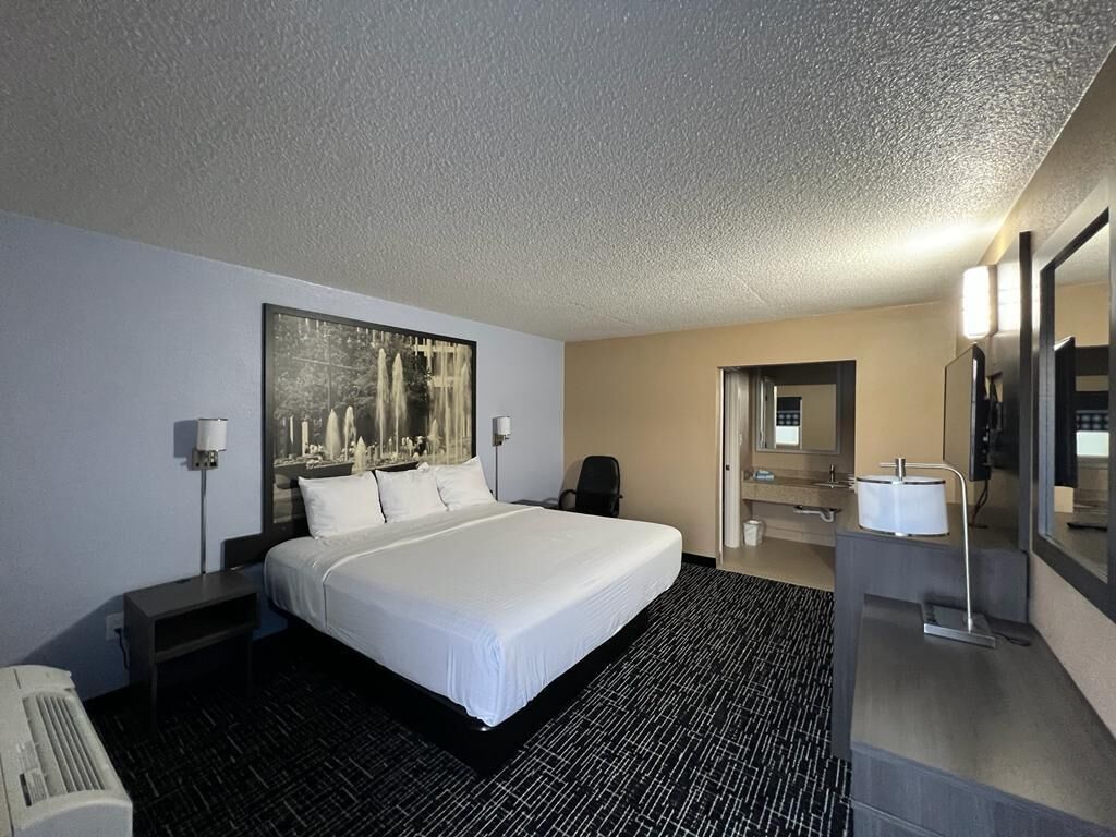 Una habitación de hotel con una cama grande, un escritorio, una silla, un baño con lavamanos y espejo, y un cuadro en la pared.