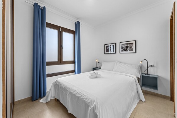 Une chambre avec un grand lit, une table de chevet, une étagère fixée au mur et une fenêtre avec des rideaux.