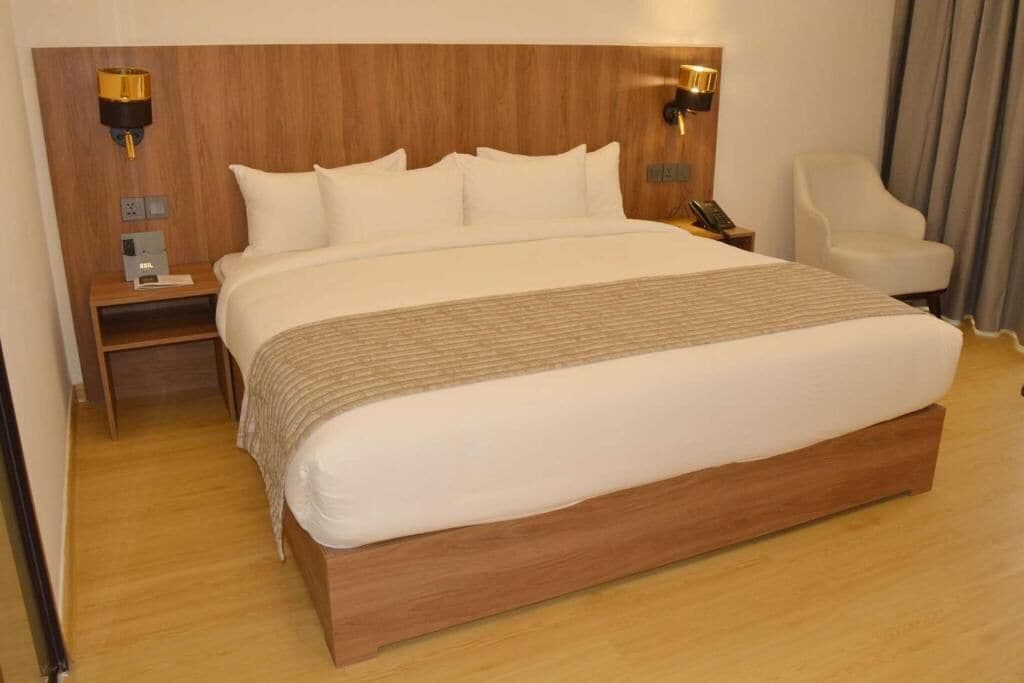 Una habitación de hotel con una cama grande, cabecera de madera, lámparas de noche, una silla y una mesita de noche.
