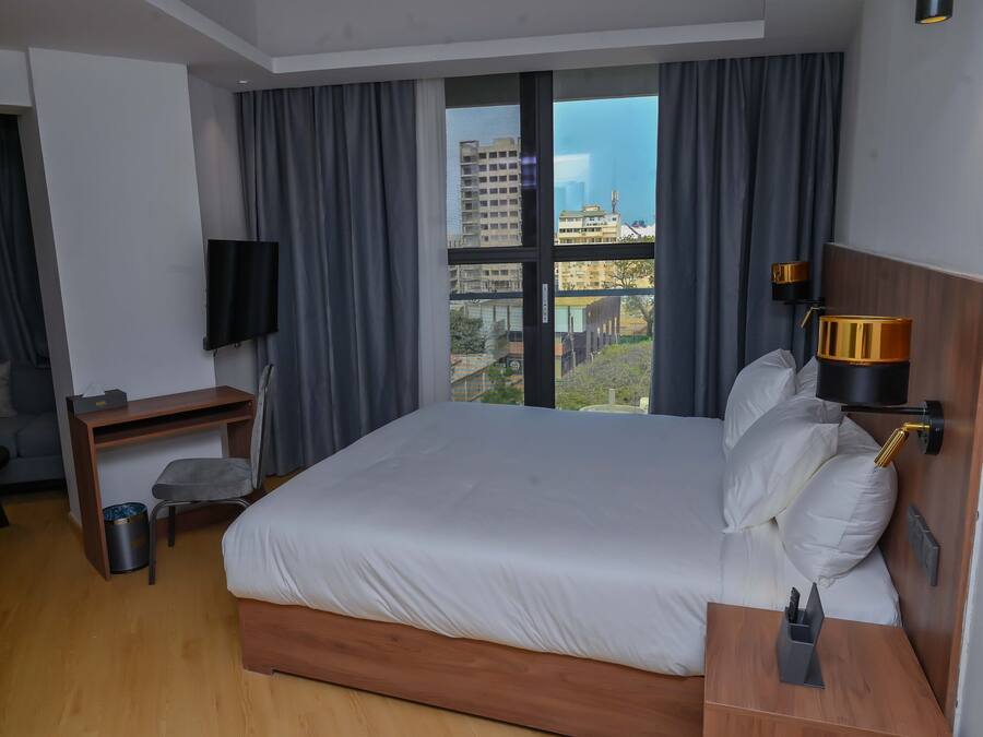 A hotel room with a large bed, a desk, a chair, a TV, a lamp, and a view of the city.