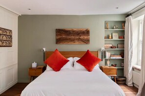 Quarto arrumado com cama grande, duas almofadas vermelhas, cabeceira de madeira, mesa de cabeceira com abajur e prateleira com diversos objetos.