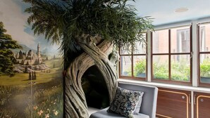 Une pièce décorée d’une fresque représentant un arbre et un château, avec un canapé agrémenté d’un coussin et de grandes fenêtres donnant sur un paysage verdoyant.