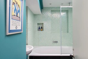Uma casa de banho com uma banheira branca, um chuveiro com divisória de vidro e uma pequena estante com produtos de higiene pessoal.