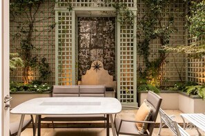 Um pátio externo com mesa e cadeiras, cercado por um painel treliçado e vegetação.