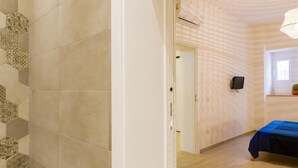 A hotel room with a wooden floor, a bed with a blue cover, a wall-mounted TV, and a door leading to another room. - LA NASSA SUITES MAZARA (Mazara del Vallo)