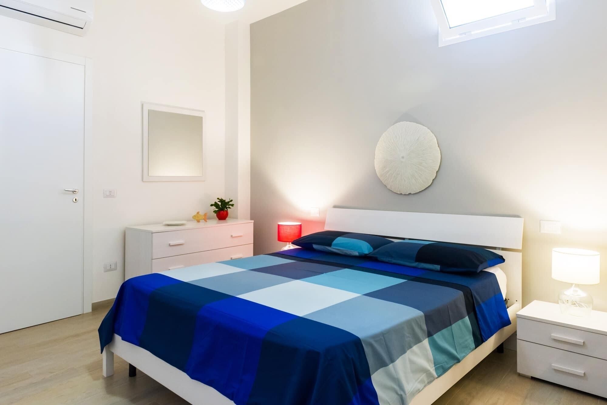 Een slaapkamer met een bed, nachtkastjes, een lamp aan de muur, een spiegel en een potplant.