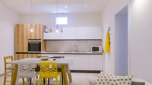 A modern kitchen with white cabinets, a microwave, and a sink. There is a dining table with chairs and a sofa with patterned pillows. - LA NASSA SUITES MAZARA (Mazara del Vallo)