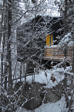 A cabin with a wooden deck and a yellow door, surrounded by snow-covered trees and rocks.
