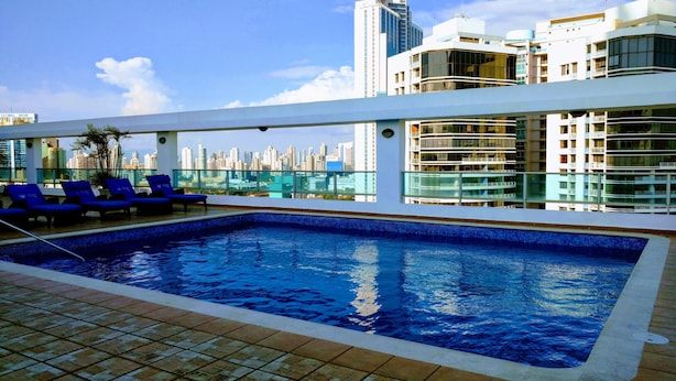 A rooftop pool with blue-tiled edges, surrounded by lounge chairs and a city skyline view.