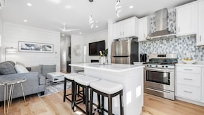 Een moderne keuken met witte kasten, een centraal eiland met barkrukken, roestvrijstalen apparatuur en een zithoek met een grijze bank en een kleine ronde tafel.