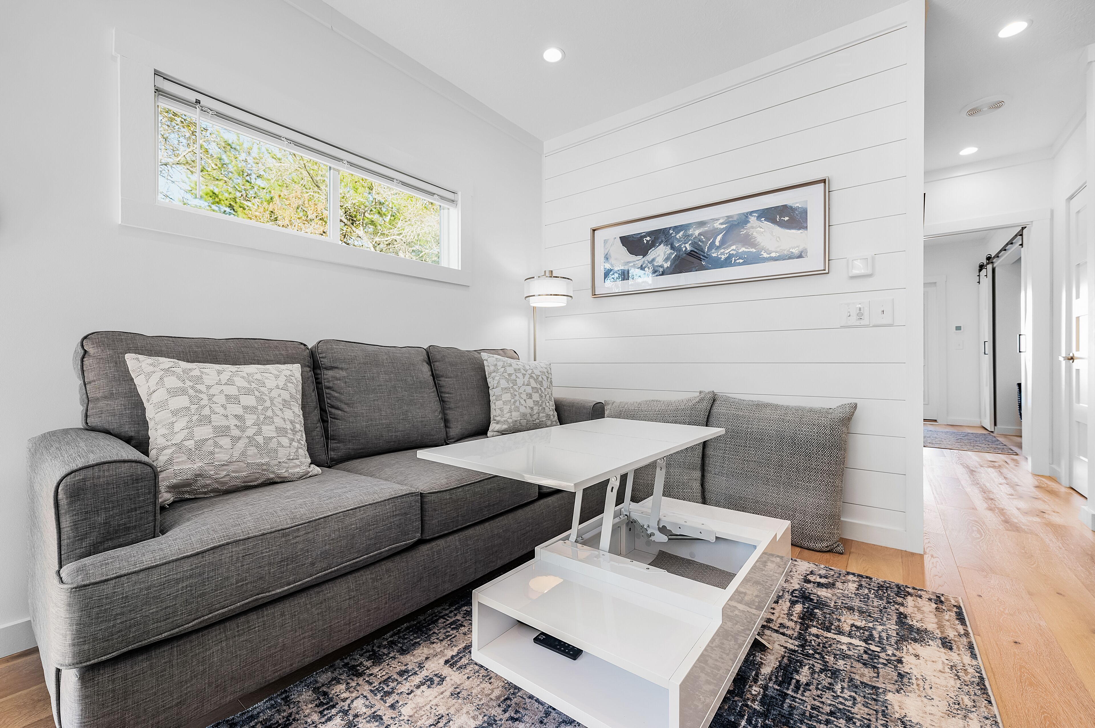 Ein modernes Wohnzimmer mit einem grauen Sofa, einem weißen Couchtisch und einem großen Fenster, das für natürliches Licht sorgt.