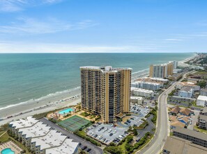 A high-rise hotel with multiple floors, a beach view, a swimming pool, and a parking lot.