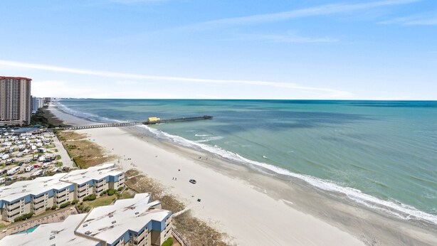 A beachside view with a pier extending into the ocean, a parking area with RVs, and a row of buildings with balconies overlooking the beach.