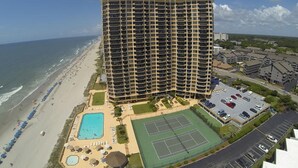 A high-rise hotel with multiple balconies, a pool, tennis courts, and a beach view.